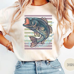 May include: Ivory t-shirt with a detailed illustration of a fish leaping from water. The fish is in shades of blue, purple, and yellow, set against a striped background of purple and green. The word "IVORY" is printed on the bottom right.