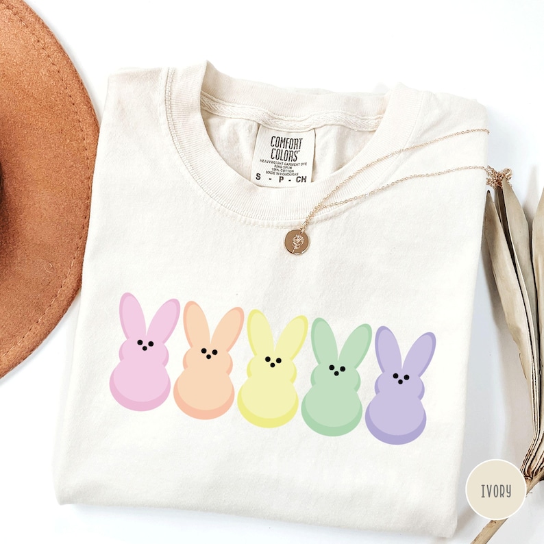 以下が含まれることがあります： アイボリー色のTシャツ。ピンク、ピーチ、イエロー、グリーン、パープルのパステルカラーのウサギの漫画のキャラクターが5匹描かれています。クルーネックと半袖。ペンダント付きのゴールドネックレスがTシャツの上にかけられています。