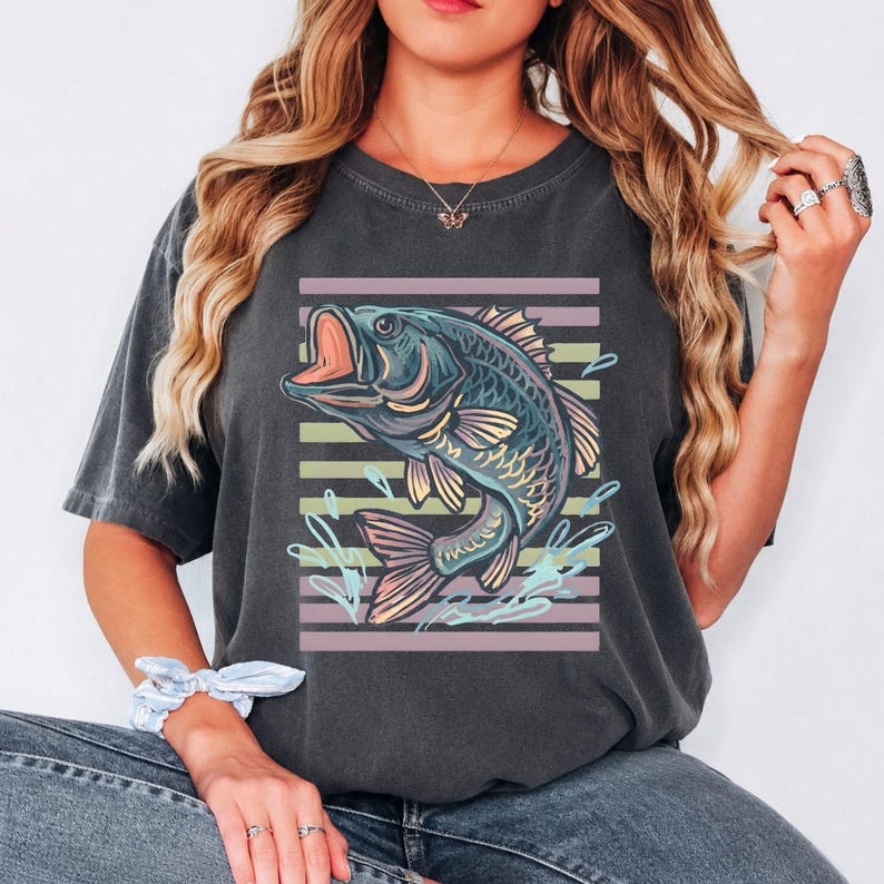 May include: Dark grey t-shirt with a detailed illustration of a largemouth bass leaping out of water. The fish is in blue, green, and yellow, set against horizontal stripes in muted colours. A fishing-themed apparel item.