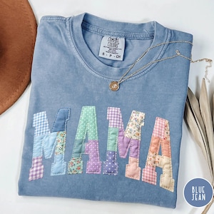 May include: Blue jean colored t-shirt with the word "MAMA" appliqued in patchwork fabric. The patchwork letters feature various patterns and colors. A gold necklace with a pendant rests on the shirt. The shirt is size small.