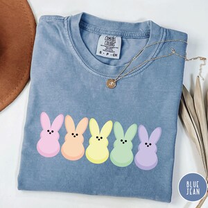 以下が含まれることがあります： パステルカラーの漫画のウサギが5匹並んだ、ブルーデニムカラーのTシャツ。Tシャツはクルーネックで半袖です。ペンダント付きのゴールドネックレスがTシャツの上にかけられています。