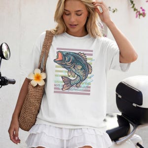 May include: White t-shirt with a detailed illustration of a fish with an open mouth, set against a backdrop of horizontal stripes in muted colours. The fish is in shades of blue, green, and pink, with water splashes around it.