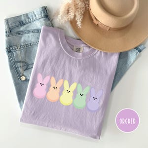 以下が含まれることがあります： パステルカラーのピンク、オレンジ、イエロー、グリーン、パープルのカラフルな漫画のウサギのキャラクターが並んだライラック色のTシャツ。右下に「ORCHID」の文字が丸く配置されています。Tシャツはブルージーンズとベージュの帽子と組み合わせています。