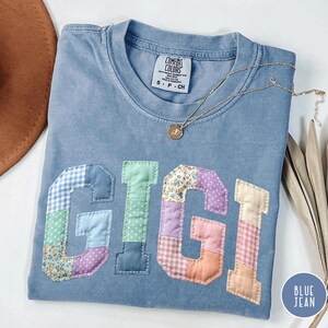 May include: A blue jean Comfort Colors t-shirt with the name "GIGI" in patchwork letters. A gold necklace with a pendant rests on the shirt. The shirt is folded, with a brown hat visible in the background.