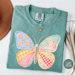 May include: Light green Comfort Colors t-shirt with a colorful patchwork butterfly design. The butterfly wings feature floral, gingham, and polka dot patterns. A silver necklace with a pendant is draped on the shirt.