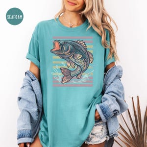 May include: Seafoam green t-shirt with a detailed illustration of a largemouth bass with an open mouth, set against a backdrop of horizontal pastel stripes. The shirt has rolled-up sleeves and a relaxed fit.