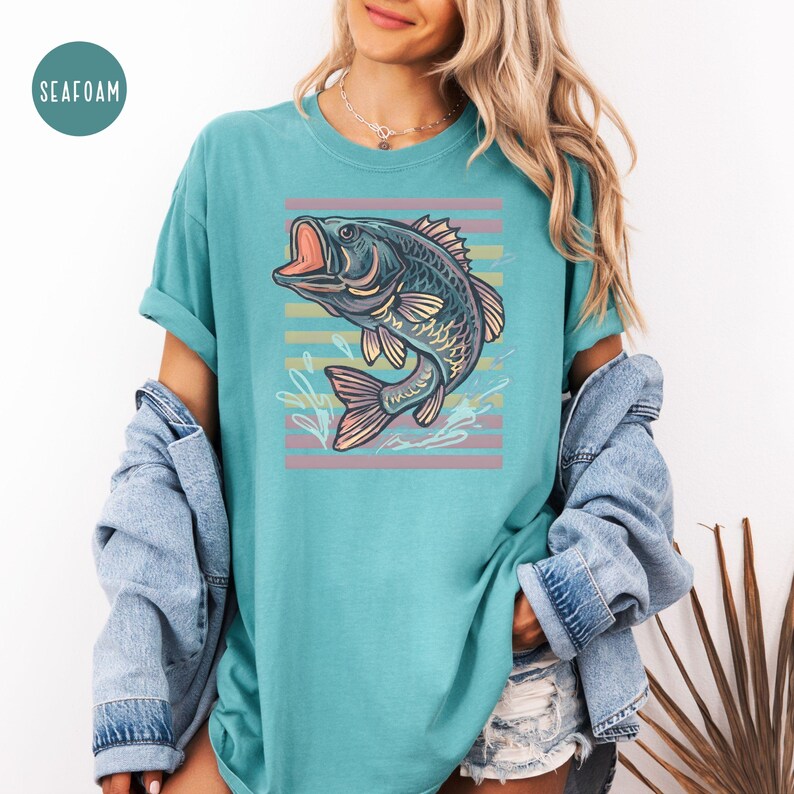May include: Seafoam green t-shirt with a detailed illustration of a largemouth bass with an open mouth, set against a backdrop of horizontal pastel stripes. The shirt has rolled-up sleeves and a relaxed fit.
