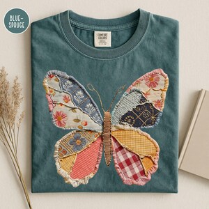 May include: Blue-spruce colored t-shirt with a patchwork butterfly design. The butterfly wings are made of various patterned fabrics in shades of pink, blue, yellow, and red. The shirt has a crew neck and a Comfort Colors tag.