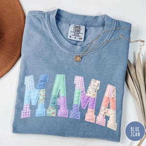 May include: Blue jean coloured t-shirt with the word "MAMA" appliquéd in patchwork fabric. The patchwork letters feature floral, polka dot, and gingham patterns. A gold necklace with a pendant rests on the shirt.