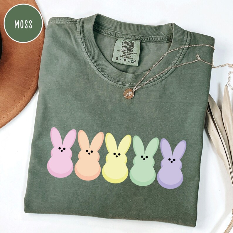 以下が含まれることがあります： モスグリーンのComfort Colors Tシャツ。パステルカラーのウサギのグラフィックが5つ並んでいます。ウサギはピンク、オレンジ、イエロー、グリーン、パープルです。ペンダント付きのゴールドネックレスがシャツの上にかけられています。
