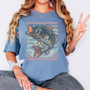 May include: A blue t-shirt with a graphic of a fish jumping out of water. The fish is in shades of blue, green, and pink, set against pastel horizontal stripes. The design includes water splashes, creating a dynamic and lively image.