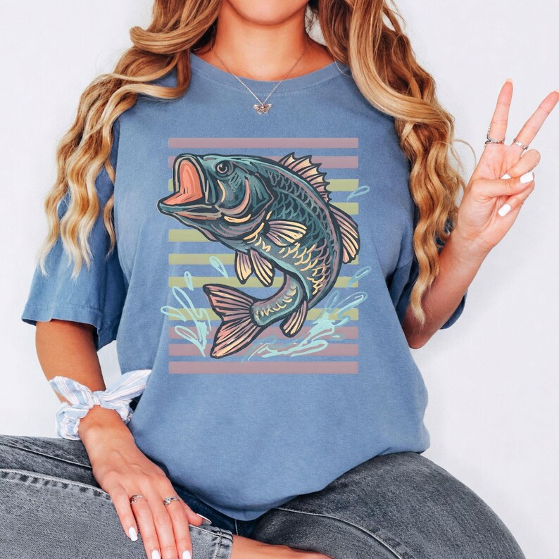 May include: A blue t-shirt with a graphic of a fish jumping out of water. The fish is in shades of blue, green, and pink, set against pastel horizontal stripes. The design includes water splashes, creating a dynamic and lively image.