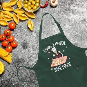 May include: Green kitchen apron with a skeleton baking a cake and the text "Having a mental bake down".