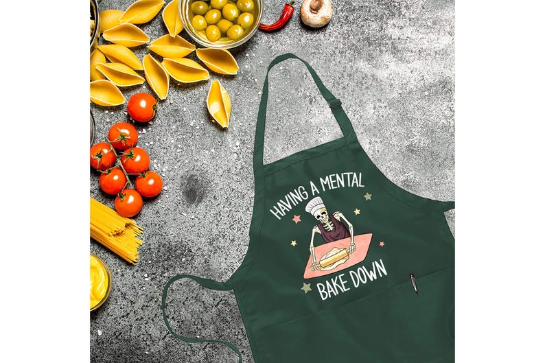 May include: Green kitchen apron with a skeleton baking a cake and the text "Having a mental bake down".