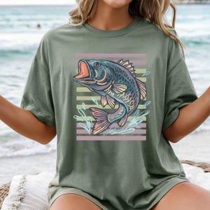 May include: Olive green t-shirt with a graphic of a fish leaping from water. The fish is blue, purple, and pink, with an open mouth. Pink and green horizontal stripes are behind the fish.