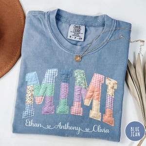 May include: Blue jean colored t-shirt with the word "MIMI" in patchwork letters. The names "Ethan", "Anthony", and "Olivia" are printed below. A gold necklace with a pendant is draped on the shirt.