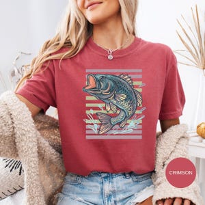 May include: Crimson-coloured t-shirt featuring a detailed illustration of a fish with an open mouth, set against a striped background. The fish is in shades of blue, green, and tan. The shirt is paired with blue denim shorts.