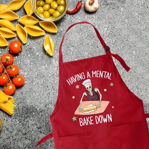 May include: Red kitchen apron with a white graphic of a skeleton baking. The text on the apron reads "Having a mental bake down."