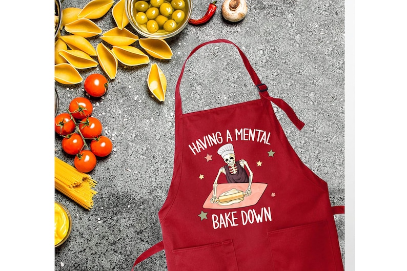 May include: Red kitchen apron with a white graphic of a skeleton baking. The text on the apron reads "Having a mental bake down."