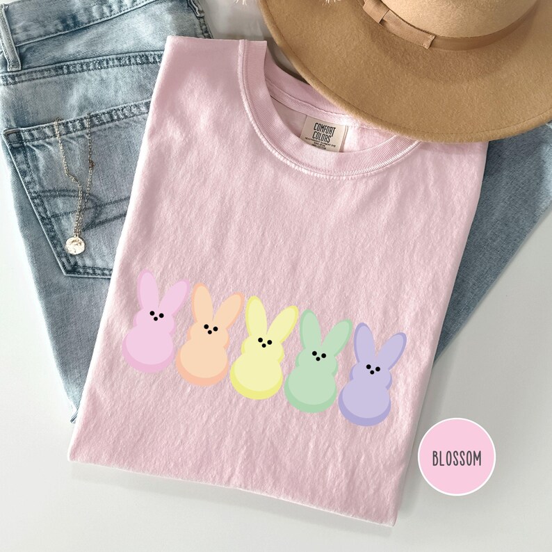 以下が含まれることがあります： パステルカラーのウサギのグラフィックが5つ並んだ、ライトピンクのComfort Colors Tシャツ。ウサギはピンク、オレンジ、イエロー、グリーン、パープルです。「BLOSSOM」という文字が入った丸いステッカーが右下にあります。