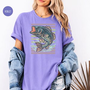 May include: A lilac-coloured t-shirt with a detailed illustration of a fish with an open mouth. The fish is in shades of blue and green, set against a backdrop of horizontal stripes in muted tones. The shirt has rolled-up sleeves.