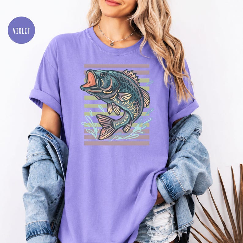 May include: A lilac-coloured t-shirt with a detailed illustration of a fish with an open mouth. The fish is in shades of blue and green, set against a backdrop of horizontal stripes in muted tones. The shirt has rolled-up sleeves.
