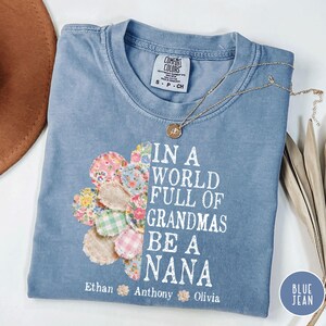 Puede incluir: Una camiseta azul vaquero Comfort Colors con el texto "IN A WORLD FULL OF GRANDMAS BE A NANA". La camiseta presenta un diseño floral y los nombres Ethan, Anthony y Olivia. Un collar dorado descansa sobre la camiseta.