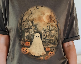 May include: A dark olive green t-shirt featuring a Halloween-themed graphic. The graphic depicts a ghost in front of a spooky house, surrounded by pumpkins, trees, and a full moon. The shirt has rolled-up sleeves.