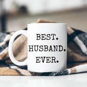 Op de afbeelding: Witte keramische mok met de tekst "BEST. HUSBAND. EVER." in zwarte, vette letters. De mok heeft een gebogen handvat en staat op een patroon deken.