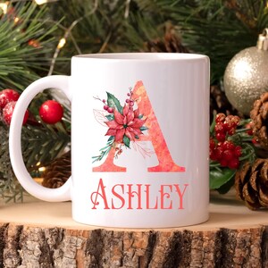 May include: White ceramic mug featuring a large, coral-colored letter "A" adorned with a floral design, including a poinsettia. The name "Ashley" is printed below in a matching coral hue. The mug is set against a festive holiday backdrop.