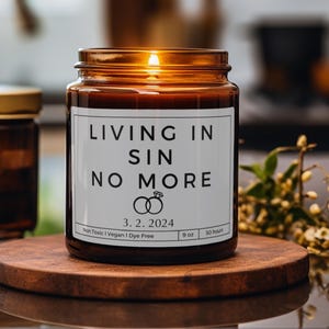 Könnte beinhalten: Eine braune Glaskerze mit einem weißen Etikett, auf dem "LIVING IN SIN NO MORE" mit einem Ringdesign und dem Datum "3.2.2024" steht. Die Kerze ist angezündet und hat ein weißes Etikett mit dem Text "Non Toxic | Vegan | Dye Free", "9 oz" und "50 hours".