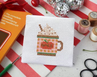 May include: A square white fabric piece featuring a cross-stitch design of a mug filled with a beverage topped with whipped cream. The mug is pink with a checkered pattern and a central orange pumpkin graphic with the word spice stitched in green.
