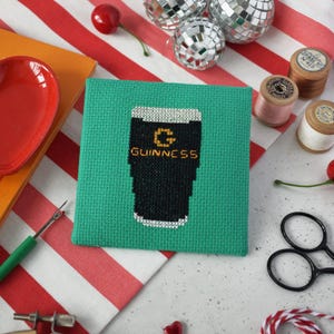 May include: A green cross-stitch coaster featuring a black and white pint of Guinness with the brand name in gold lettering. The coaster is square and sits on a red and white striped surface.