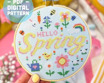 May include: A blue and white embroidery hoop with a cross stitch design that says "Hello Spring" in yellow and pink lettering. The design also includes a rainbow, a watering can, a bird, and flowers.