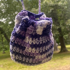 Könnte beinhalten: Eine kleine, handgefertigte Häkeltasche in Lila- und Weißtönen. Die Kordelzugtasche wird von einer passenden lila Kordel gehalten. Das Häkelmuster ist sichtbar, und die Tasche scheint aus weichem Garn zu bestehen.