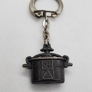 May include: A vintage metal keychain featuring a small, black pot with the word "SEB" embossed on the front. The keychain has a silver ring and chain.