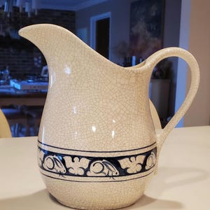 May include: A white ceramic pitcher with a crackled glaze and a blue and white floral design featuring rabbits. The pitcher has a wide mouth and a handle.