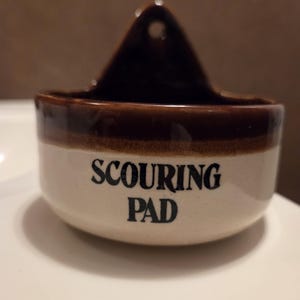 May include: A ceramic scouring pad holder with a brown rim and a brown triangular hanger. The holder is off-white with the words "SCOURING PAD" in black lettering. The holder is designed to be mounted on a wall.
