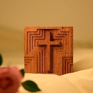 May include: A wooden cross carved into a square block of wood. The cross is raised and the background is carved with a geometric pattern. The block is sitting on a beige fabric surface.