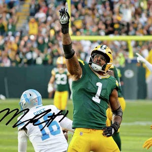 May include: A football player in a green and yellow uniform with the number 1 on the jersey raises his arm in celebration. The player wears a helmet and gloves. The background shows a crowd of people in a stadium.
