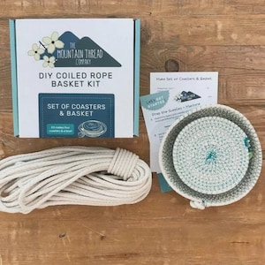 May include: A DIY coiled rope basket kit in a white box labeled The Mountain Thread Company, including a long coil of off-white rope, a set of instructions, and a finished round coiled rope bowl with green stitching details on the rim.