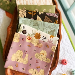 May include: A stack of six folded fabric squares featuring autumn-themed patterns including pumpkins, sunflowers, leaves, and coffee cups in shades of mauve, cream, sage green, and dark brown, resting on a wooden tray with the text Parker on the Porch.