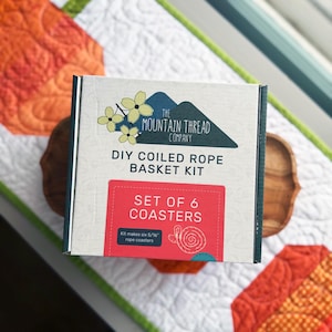 May include: A square cardboard box for The Mountain Thread Company DIY Coiled Rope Basket Kit. The label features a mountain graphic, yellow flowers, and a red rectangle stating Set of 6 Coasters, noting it makes six 5/16 inch rope coasters.