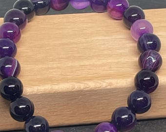 May include: A beaded bracelet featuring a variety of purple and violet gemstones. The bracelet is composed of round, polished beads in shades ranging from deep violet to light lavender. The beads are strung together, creating a circular design.