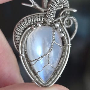 Puede incluir: Colgante de plata hecho a mano con una piedra lunar iridiscente en forma de pera. La piedra está encerrada en un intrincado trabajo de alambre con un diseño de vid torcida. El colgante está suspendido de una argolla en bucle. El texto "Crystal Story" es visible.