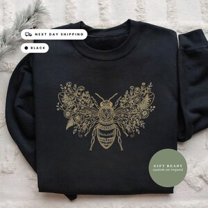 May include: A black crewneck sweatshirt featuring a gold-colored graphic print of a bee. The bee's wings are intricately detailed with various floral and botanical illustrations. The sweatshirt has long sleeves and a ribbed collar.