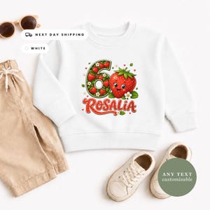 May include: A white long-sleeve sweatshirt featuring a graphic of the number six filled with small strawberries, a large smiling strawberry character, and the name Rosalia in red script lettering. The design includes small white flowers.