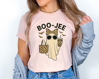 May include: A light pink t-shirt featuring a graphic of a ghost wearing sunglasses and a leopard print bow. The ghost holds a leopard print tote bag and a drink cup. The text BOO-JEE is printed above the ghost, surrounded by small bats.
