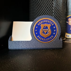 May include: A blue and gold business card holder with the words "Undercover Association" and a gold shield with a blue fedora on top.
