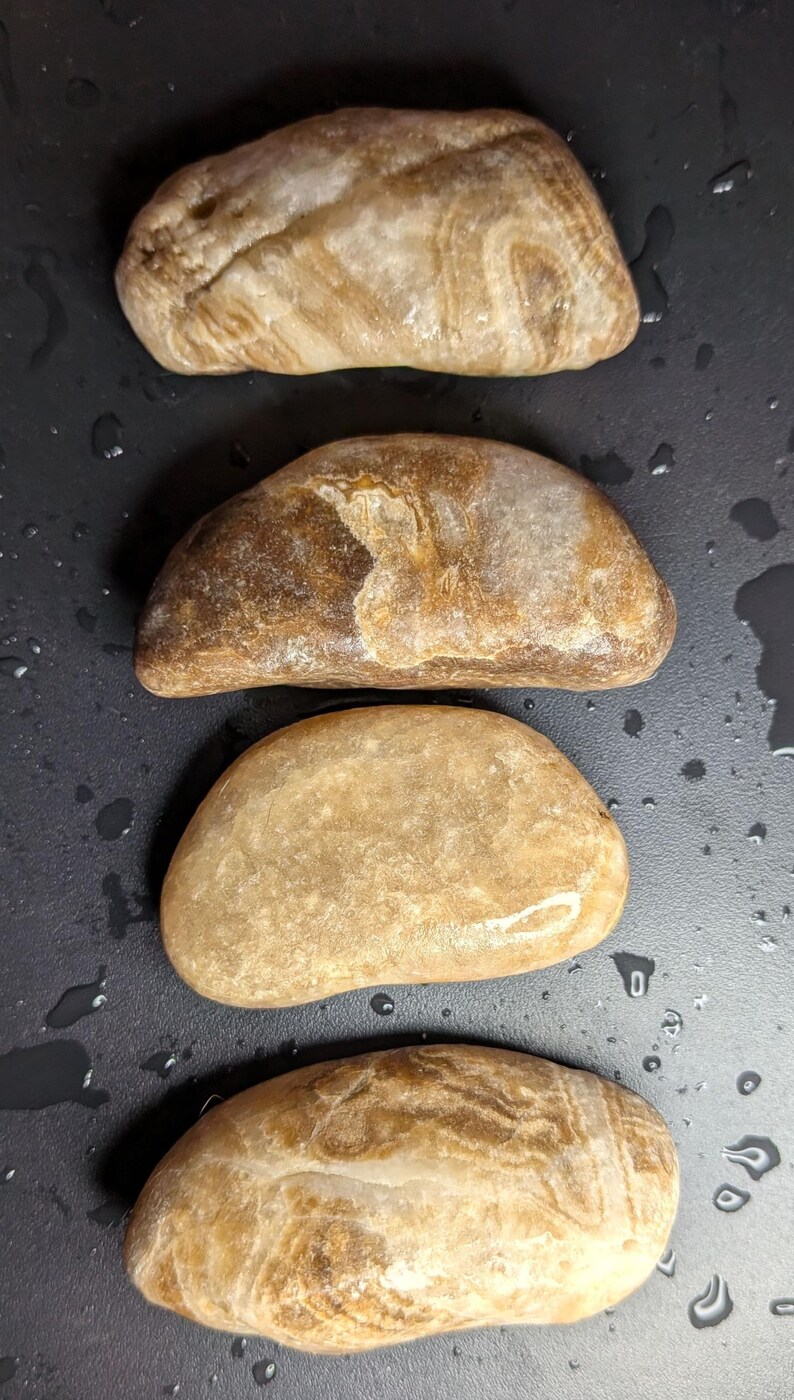 Puede incluir: Cuatro piedras lisas de forma ovalada en tonos beige y marr&oacute;n. Las piedras tienen patrones y texturas &uacute;nicos, con algunas que muestran estr&iacute;as en capas. Las piedras est&aacute;n dispuestas verticalmente sobre un fondo oscuro.